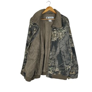 May include: A camouflage jacket with a brown interior and a collar. The jacket is suspended on a wooden hanger. The Columbia logo is visible on the label. The jacket's exterior features a mix of green, brown, and grey tones.