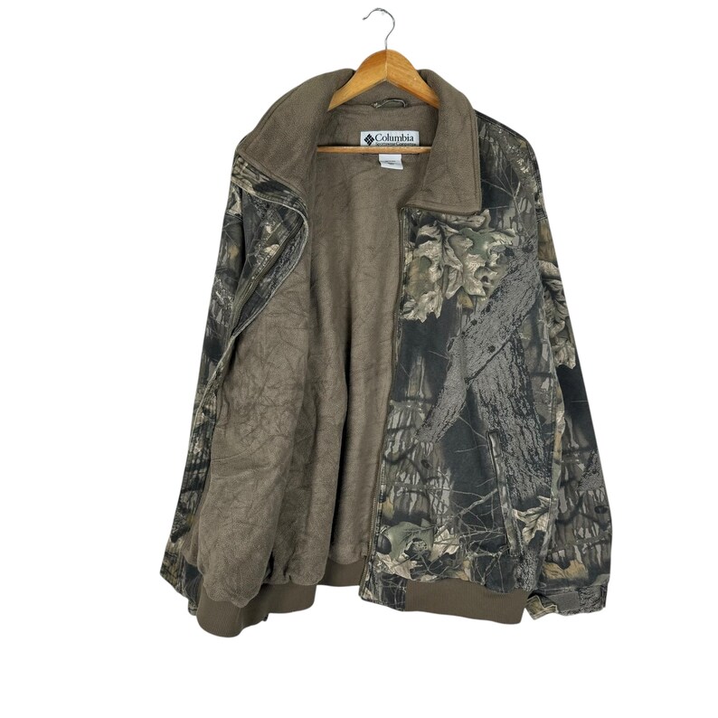 May include: A camouflage jacket with a brown interior and a collar. The jacket is suspended on a wooden hanger. The Columbia logo is visible on the label. The jacket's exterior features a mix of green, brown, and grey tones.