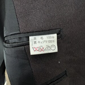 Op de afbeelding: Close-up van een donkerbruine blazer met een zwarte voering en een klein wit label. Het label heeft Japanse tekst en wassymbolen. De stof van de blazer lijkt getextureerd.