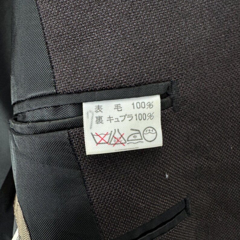 Op de afbeelding: Close-up van een donkerbruine blazer met een zwarte voering en een klein wit label. Het label heeft Japanse tekst en wassymbolen. De stof van de blazer lijkt getextureerd.