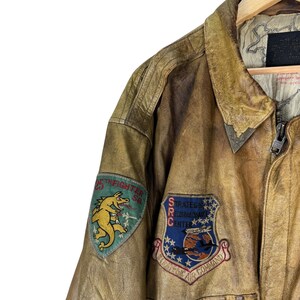May include: Close-up of a vintage brown leather jacket with patches. The jacket features a zipper closure and a collared neckline. Patches include a dragon and text that reads "25th Fighter Sq" and "Strategic Reconnaissance Center".