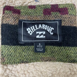 May include: A black and white label with the word "Billabong" in a stylised font. The label has a small white wave logo and the text "S P CH MADE IN CHINA". The label is attached to a green, brown and red plaid fabric.