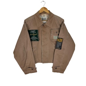 Peut inclure: Veste de couleur beige avec un col et des boutons. La veste comporte plusieurs poches et des écussons avec du texte, dont "Finest Fabric" et "426". Elle est suspendue à un cintre en bois.