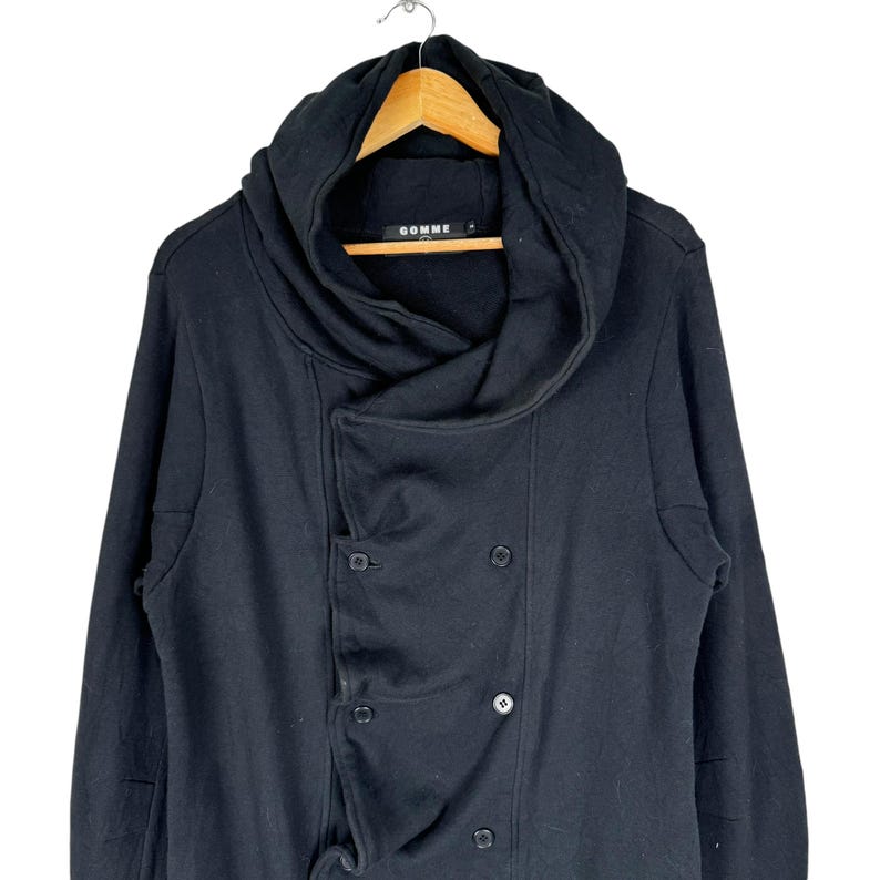 May include: Black hooded long-sleeved jacket with a double-breasted closure and a large, draped collar. The jacket has the word "Comme" printed on the label.