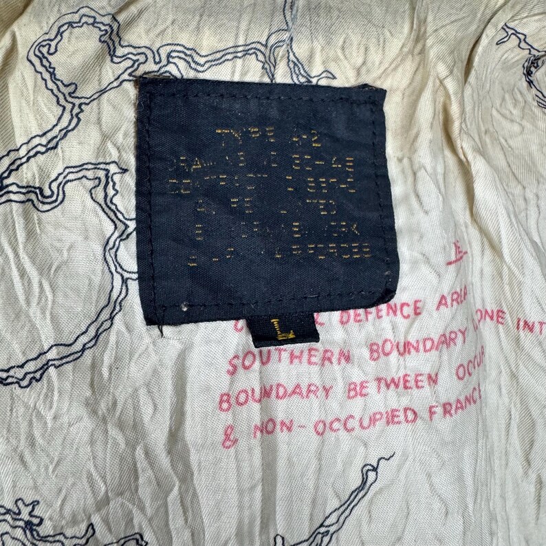May include: Close-up of a vintage jacket's interior, featuring a dark blue label with text and a size "L". The lining is off-white with a map-like design in black. Red text is printed on the lining.