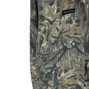 May include: A camouflage jacket with a full zip and a small pocket. The jacket features a pattern of green, brown, and tan leaves. The Columbia Sportswear Company logo is visible on the upper right side.
