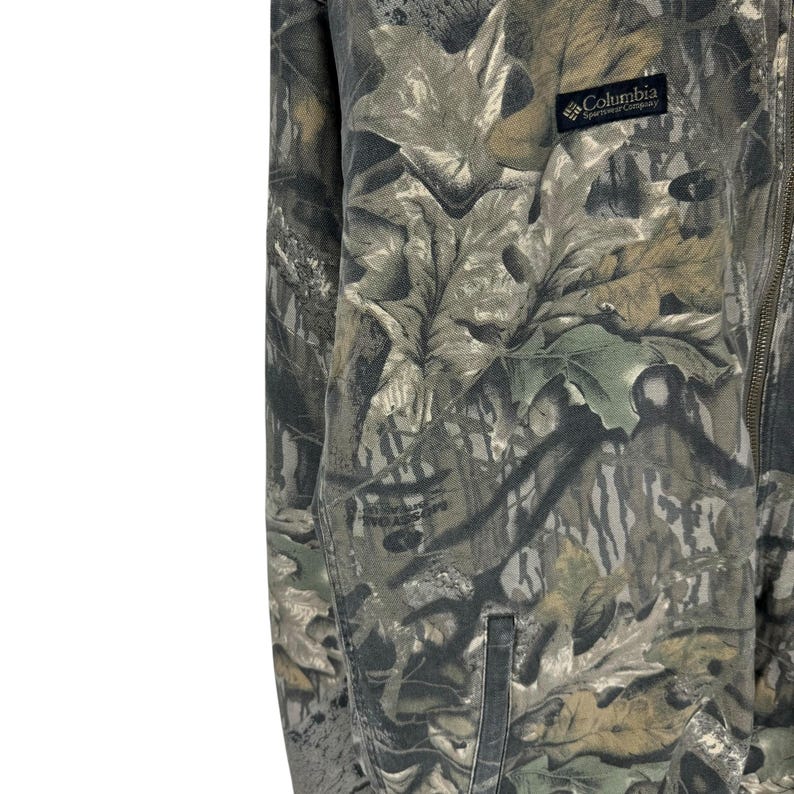 May include: A camouflage jacket with a full zip and a small pocket. The jacket features a pattern of green, brown, and tan leaves. The Columbia Sportswear Company logo is visible on the upper right side.