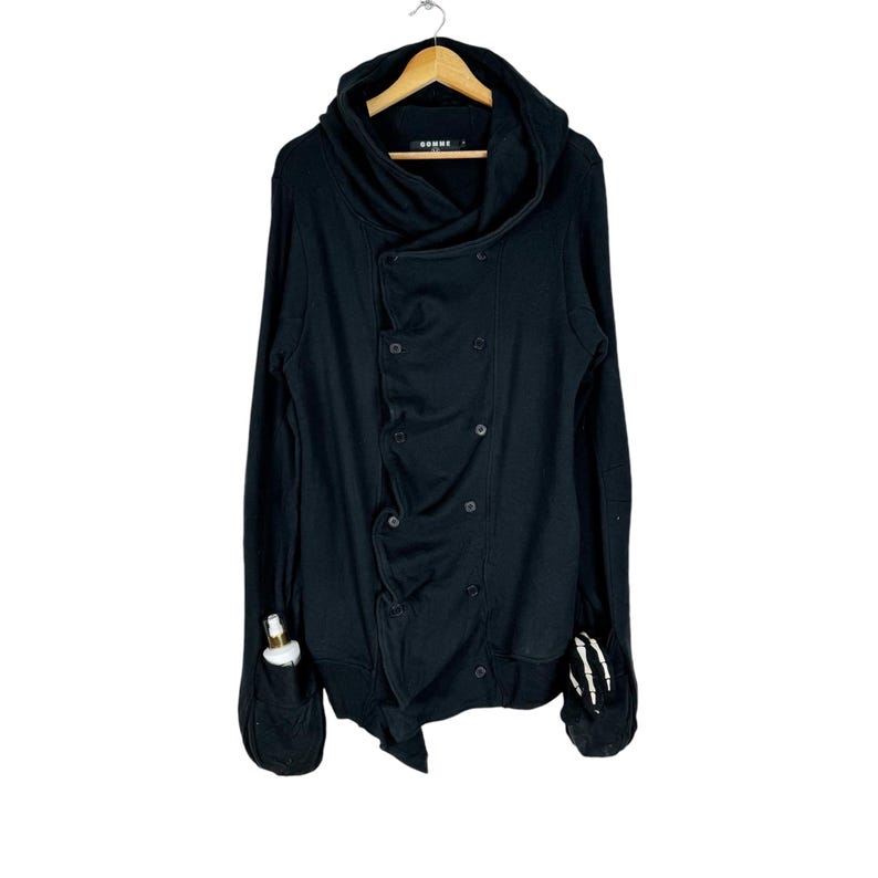 May include: Black hooded sweatshirt with a double-breasted button closure and a long, asymmetrical hem. The sweatshirt has a skull graphic on the right sleeve and the word "DOMME" printed on the inside of the hood.