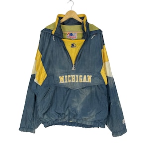 Puede incluir: Una chaqueta cortavientos de la marca Starter, de color azul y amarillo, con la palabra "MICHIGAN" en letras amarillas en la parte delantera. La chaqueta tiene una cremallera y una capucha.