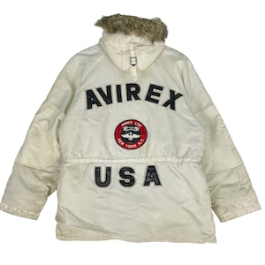 Peut inclure: Une veste Avirex blanche avec une capuche en fausse fourrure. La veste est ornée d'un logo brodé noir qui dit "Avirex" et d'un patch qui dit "Avirex Ltd. New York N.Y.". La veste a également un patch brodé noir qui dit "USA".