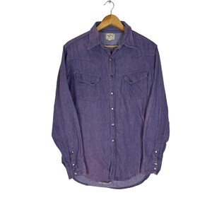 May include: A purple denim long-sleeve shirt with a button-down collar, two chest pockets, and a single button cuff. The shirt has a faded look and is made of a soft, comfortable fabric.