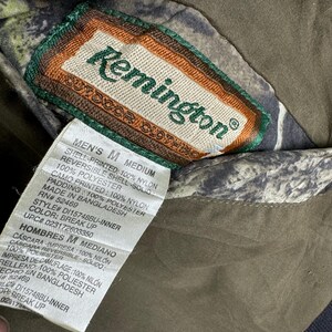 May include: A brown and green camouflage jacket with a label that reads "Remington Outdoor Clothing" and "Men's M Medium". The label lists the materials used in the jacket, including nylon and polyester. The label also states that the jacket was made in Bangladesh.