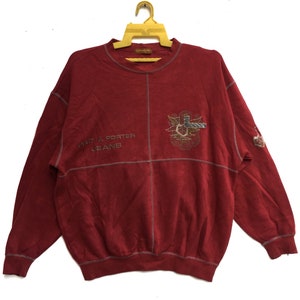 Könnte beinhalten: Roter Langarm-Pullover mit einem "Pret a Porter Jeans" Logo und einem gestickten Design auf der Vorderseite.
