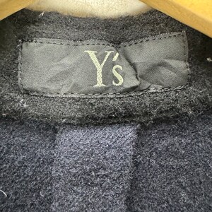 K&ouml;nnte beinhalten: Nahaufnahme eines schwarzen Etiketts mit dem Text "Y's" in wei&szlig;er Stickerei. Das Etikett ist an einem dunkelblauen Stoffkleidungsst&uuml;ck befestigt.