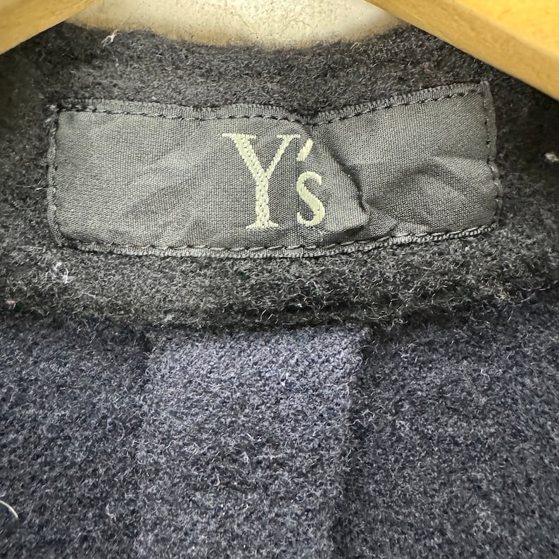 K&ouml;nnte beinhalten: Nahaufnahme eines schwarzen Etiketts mit dem Text "Y's" in wei&szlig;er Stickerei. Das Etikett ist an einem dunkelblauen Stoffkleidungsst&uuml;ck befestigt.