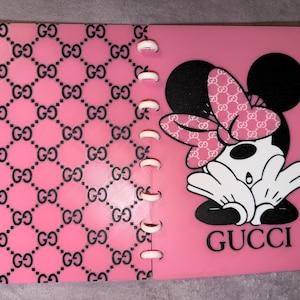May include: A pink notebook with a repeating black interlocking GG logo pattern on the left cover. The right cover features a black and white cartoon mouse character with a pink patterned bow and the word GUCCI printed in black at the bottom.