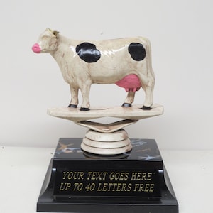 May include: A black and white cow trophy with pink accents stands on a black base with a gold inscription that reads "YOUR TEXT GOES HERE UP TO 40 LETTERS FREE".