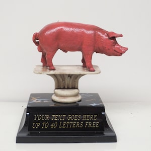 May include: A red pig trophy with a black base and a white marble-like pedestal. The base has the text "YOUR TEXT GOES HERE UP TO 40 LETTERS FREE".