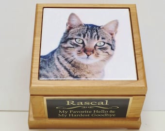 May include: A wooden pet urn with a personalised plaque that reads "Rascal, My Favourite Hello & My Hardest Goodbye". The urn has a ceramic tile on the front with a photo of a tabby cat.