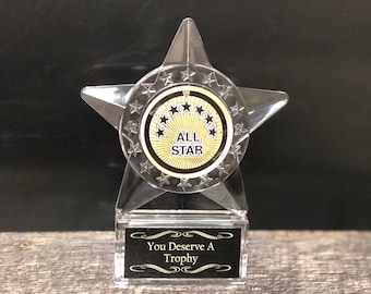May include: A clear acrylic trophy shaped like a star with a black and gold circular medallion that says "ALL STAR" and five black stars. The base of the trophy is black with gold trim and the text "You Deserve A Trophy".