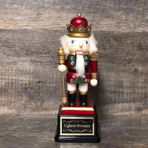 May include: A red and green wooden nutcracker with a gold crown and a white beard. The nutcracker is holding a gold staff and is standing on a black base with the words "Ugliest Sweater" written on it.