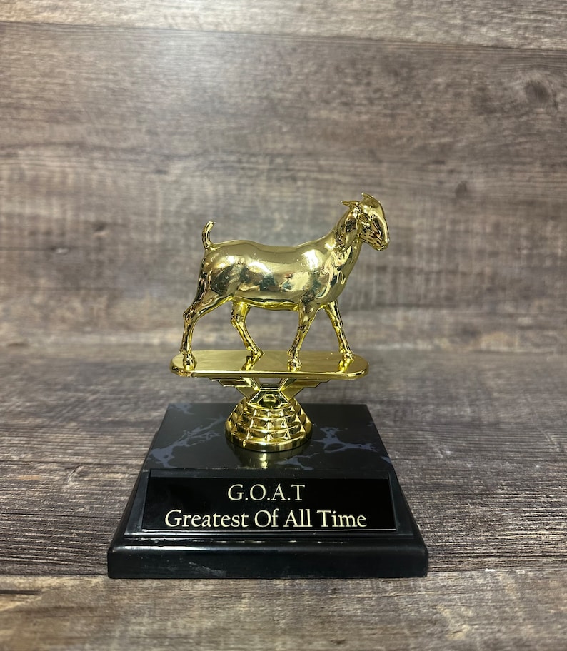 May include: A gold-colored trophy featuring a goat figurine standing on a black marble base. The base has a gold-colored plate with the text "G.O.A.T. Greatest Of All Time".