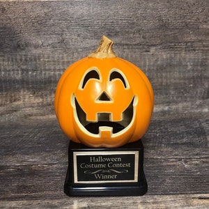 May include: A Halloween costume contest winner trophy featuring a smiling orange pumpkin with a black base and a gold plaque that reads "Halloween Costume Contest Winner".