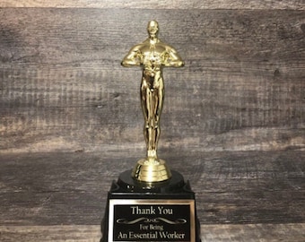 May include: Gold-colored trophy with a black base. The trophy features a standing figure with arms outstretched. The base has a black plaque with the text "Thank You For Being An Essential Worker".