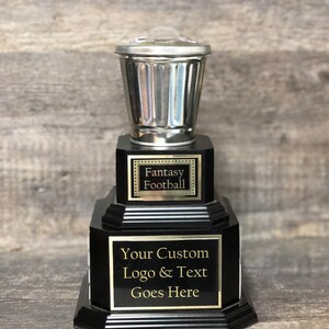 May include: A black and silver trophy with a trash can shaped top. The trophy has a black base with a silver plaque that says "Fantasy Football". The bottom plaque says "Your Custom Logo & Text Goes Here".