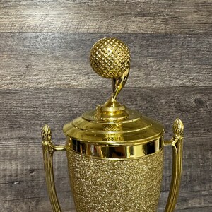 Może przedstawiać: Złoty puchar golfowy z fakturowanym wykończeniem. Puchar ma na szczycie piłkę golfową na trzonku.