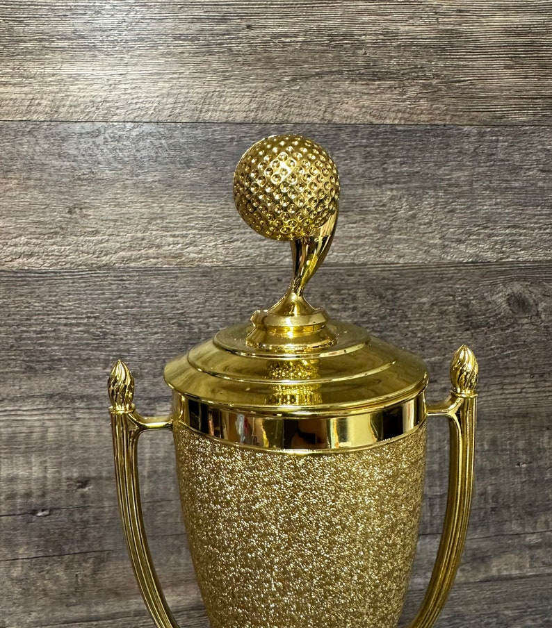 Może przedstawiać: Złoty puchar golfowy z fakturowanym wykończeniem. Puchar ma na szczycie piłkę golfową na trzonku.