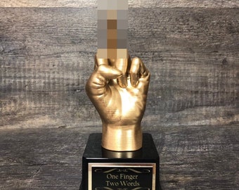 May include: A gold-colored trophy with a hand giving the middle finger. The base of the trophy is black with a gold plaque that reads "One Finger Two Words".