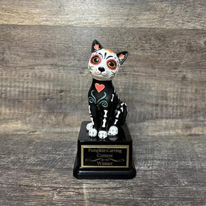 May include: Black and white cat figurine with a sugar skull design, sitting on a black base with a gold plaque that reads "Pumpkin Carving Contest Winner".