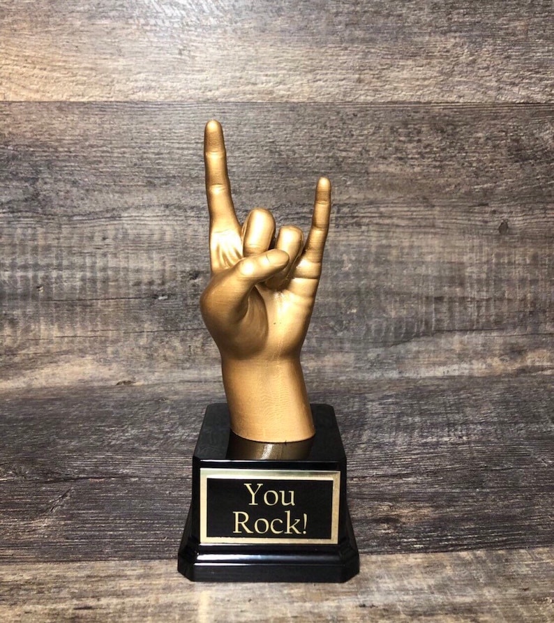 Puede incluir: Una estatua de mano dorada en el gesto de rock and roll, montada sobre una base negra. La base tiene una placa con borde dorado con las palabras "You Rock!" en letras doradas. El fondo es una superficie de madera.
