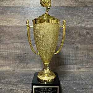 Może przedstawiać: Złoty puchar golfowy z czarną podstawą. Puchar ma na szczycie piłkę golfową i tabliczkę z napisem "Hole In One" z imieniem "John Smith" i datą "6/10/2022".