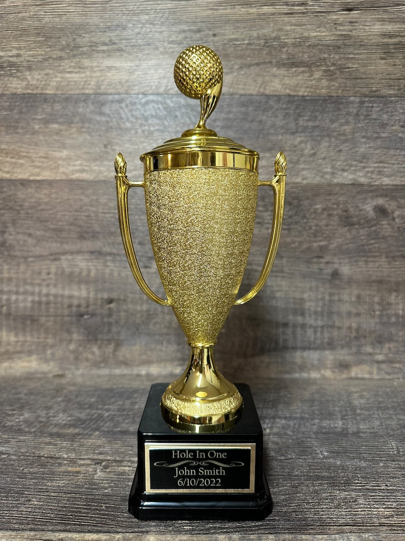 Może przedstawiać: Złoty puchar golfowy z czarną podstawą. Puchar ma na szczycie piłkę golfową i tabliczkę z napisem "Hole In One" z imieniem "John Smith" i datą "6/10/2022".