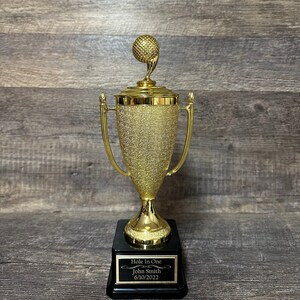 Może przedstawiać: Złoty puchar golfowy z czarną podstawą. Podstawa ma tabliczkę z napisem "Hole In One" z imieniem "John Smith" i datą "6/10/2022".