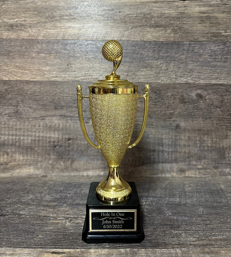 Może przedstawiać: Złoty puchar golfowy z czarną podstawą. Podstawa ma tabliczkę z napisem "Hole In One" z imieniem "John Smith" i datą "6/10/2022".