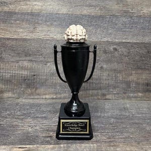 Puede incluir: Un trofeo negro con una parte superior en forma de cerebro. La base del trofeo tiene una placa dorada que dice "Knowledge Bowl Champion".