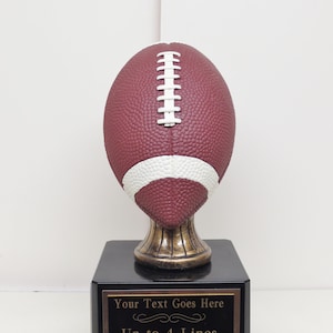 Puede incluir: Un trofeo de fútbol marrón y blanco con una base negra. La base tiene una placa dorada con el texto "Your Text Goes Here" y "Up to 4 Lines Engraved Free".