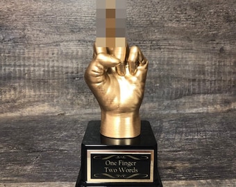 May include: A gold-colored hand sculpture making the middle finger gesture. The hand is mounted on a black base with a gold plaque that reads "One Finger Two Words".