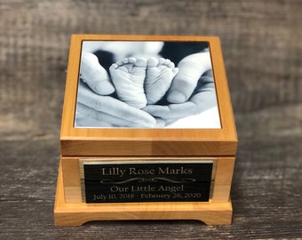 Puede incluir: Una caja de recuerdo de madera con una foto en blanco y negro de los pies de un bebé en la tapa. La caja tiene una placa dorada con la inscripción "Lilly Rose Marks Our Little Angel July 10, 2018 - February 26, 2020".