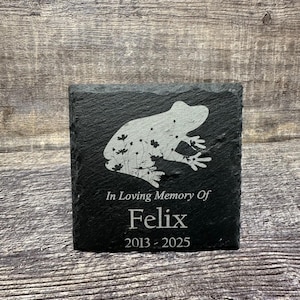 Puede incluir: Piedra conmemorativa de pizarra negra con una silueta blanca de una rana y flores. El texto "In Loving Memory Of Felix 2013 - 2025" está grabado en la piedra.