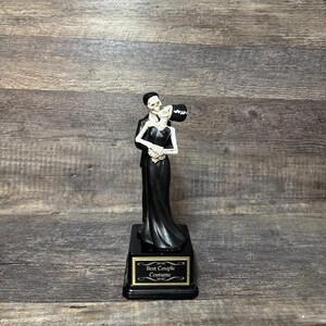 May include: A black and white skeletal couple figurine holding each other, wearing black clothing. The figurine is on a black base with a gold plaque that reads "Best Couple Costume".