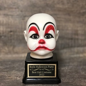 May include: A black and gold trophy with a white porcelain clown head on top. The trophy has a plaque that reads "Boyle Halloween Party Best Girl Costume 2022".
