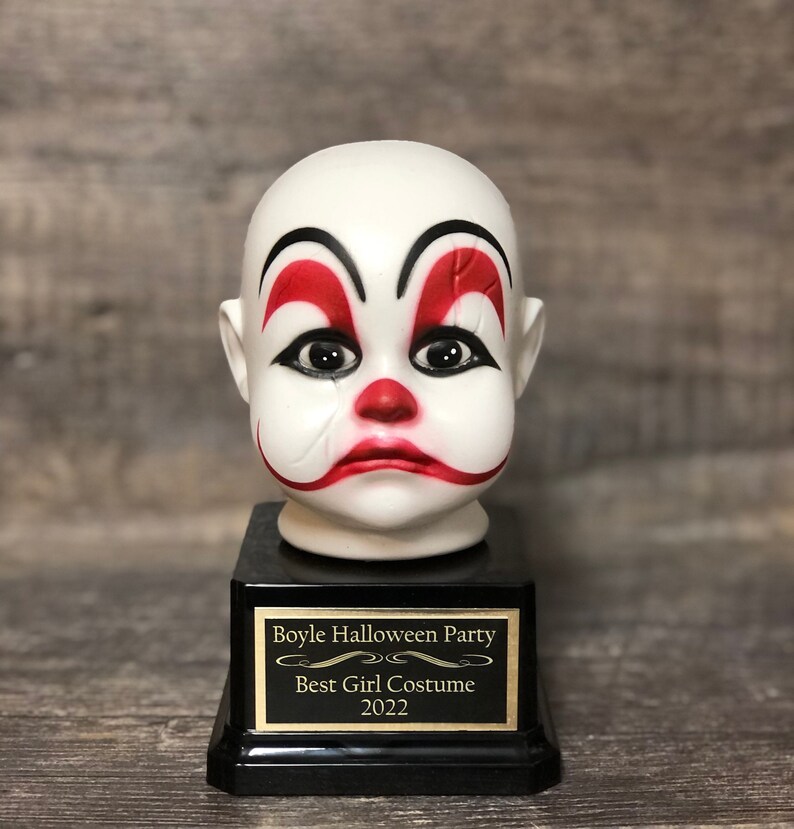 May include: A black and gold trophy with a white porcelain clown head on top. The trophy has a plaque that reads "Boyle Halloween Party Best Girl Costume 2022".