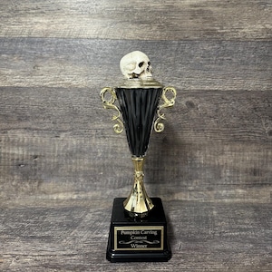 May include: A trophy featuring a realistic off-white human skull topper mounted on a black fluted cup with gold-toned handles and base. The black square pedestal base displays a gold plaque with the text Pumpkin Carving Contest Winner.