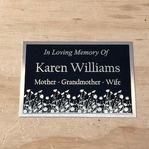 Può includere: Una placca d'argento con sfondo nero e lettere bianche. La placca recita "In Loving Memory Of Karen Williams Mother - Grandmother - Wife". La parte inferiore della placca presenta un motivo floreale bianco.