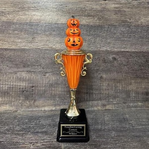 May include: A gold trophy with a black base and a pumpkin carving contest winner inscription. The trophy features three stacked orange pumpkins with carved faces.
