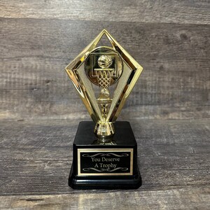 May include: A gold basketball trophy with a black base. The trophy features a basketball going through a hoop. The base has a black plaque with the text "You Deserve A Trophy".